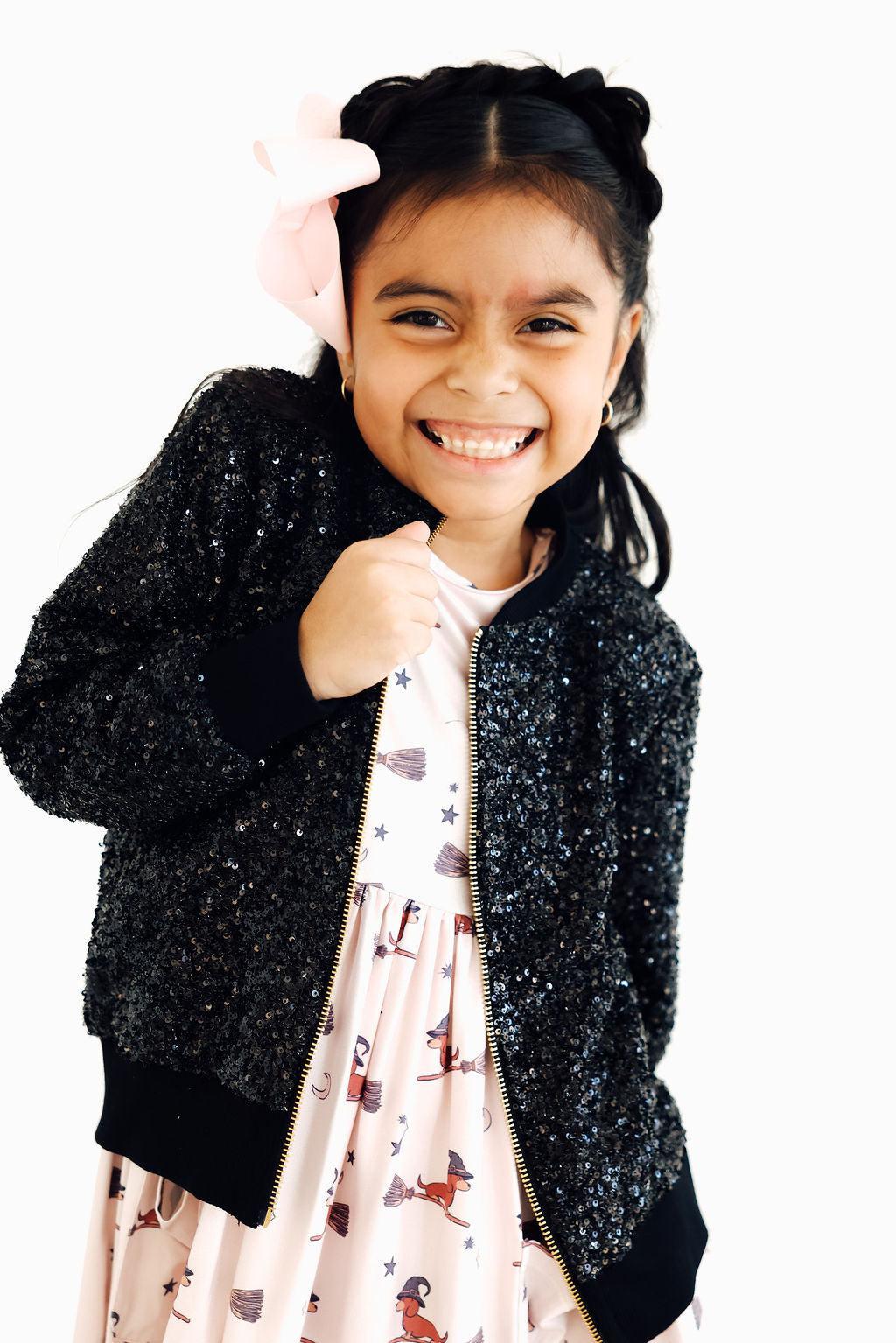 Girl smiling as she holds the black sequin jacket open over a light pink dress printed with witches, brooms and stars, with a pink bow in her hair.