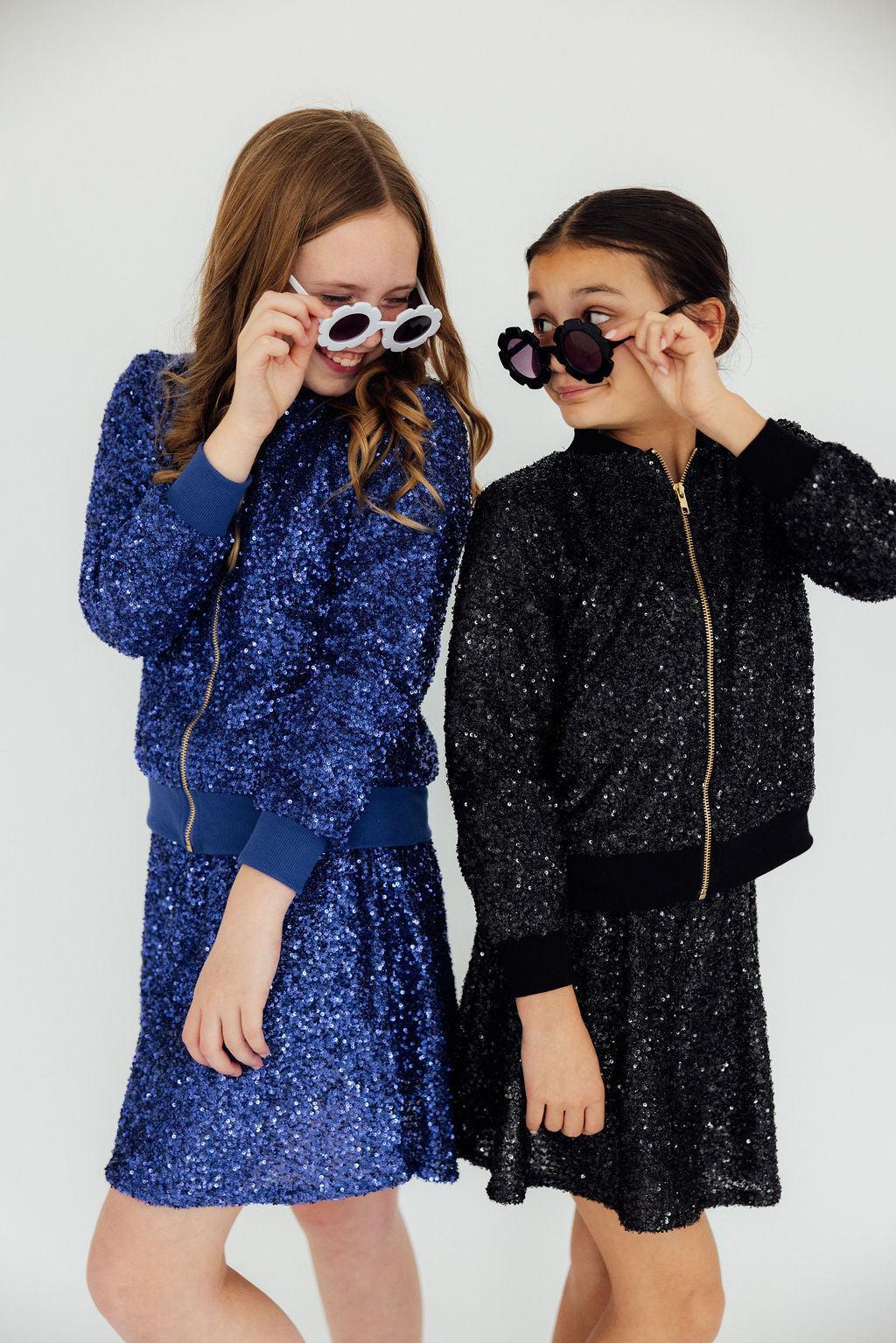 Two girls side by side in Mila & Rose sequin bomber jackets, one royal blue and one black, each with a matching sequin skirt.