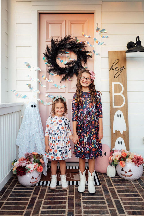 Girl wearing the Boo Crew twirl dress on a porch beside another child, in front of a Halloween door display.
