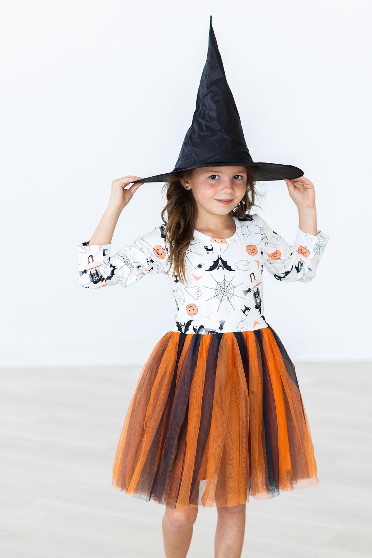 Front view of the Boo-tiful Halloween L/S Tutu Dress, full length, with a girl holding the brim of a black witch hat prop she is wearing; printed white bodice and orange and black layered tulle skirt.