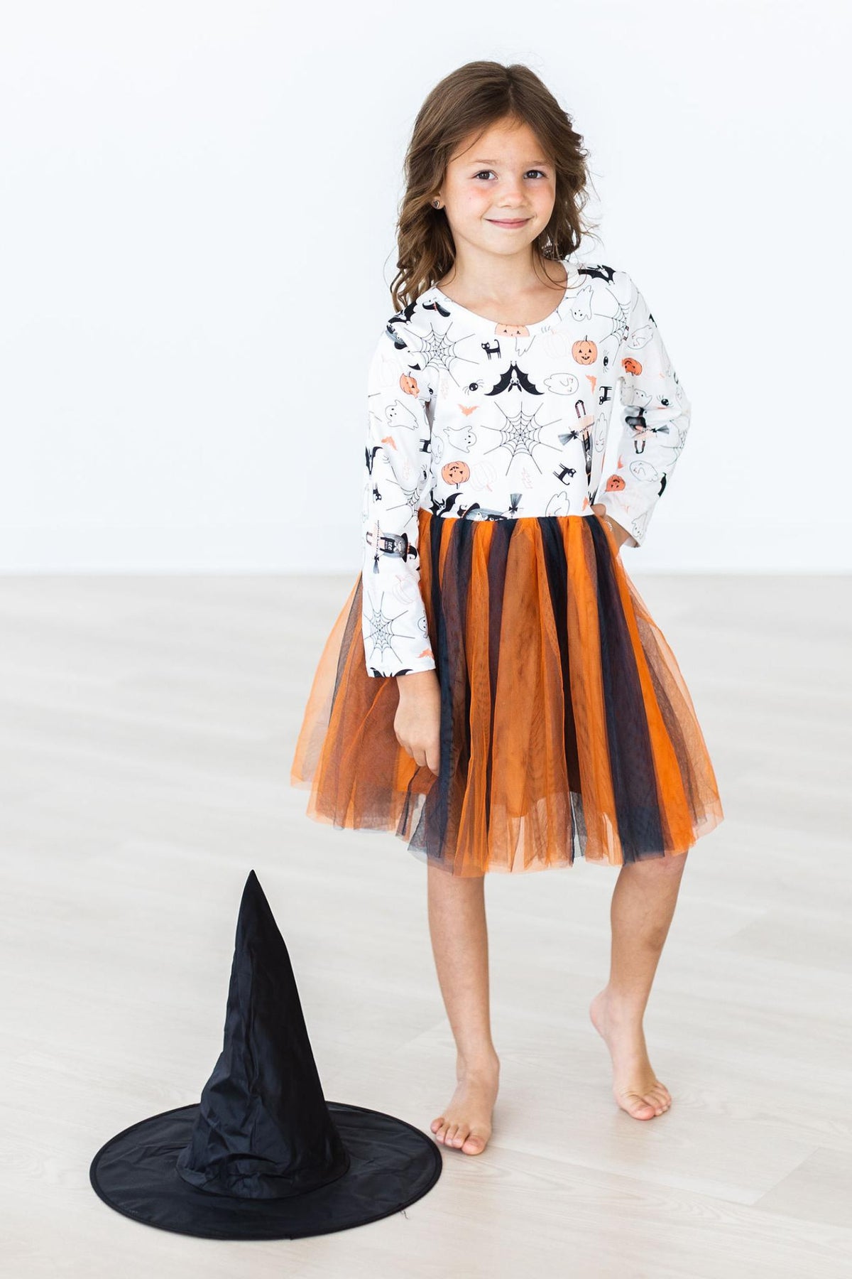 Girl standing in the Boo-tiful Halloween long-sleeve tutu dress with the layered orange and black tulle skirt flared; the black witch hat is a photo prop resting on the floor beside her.