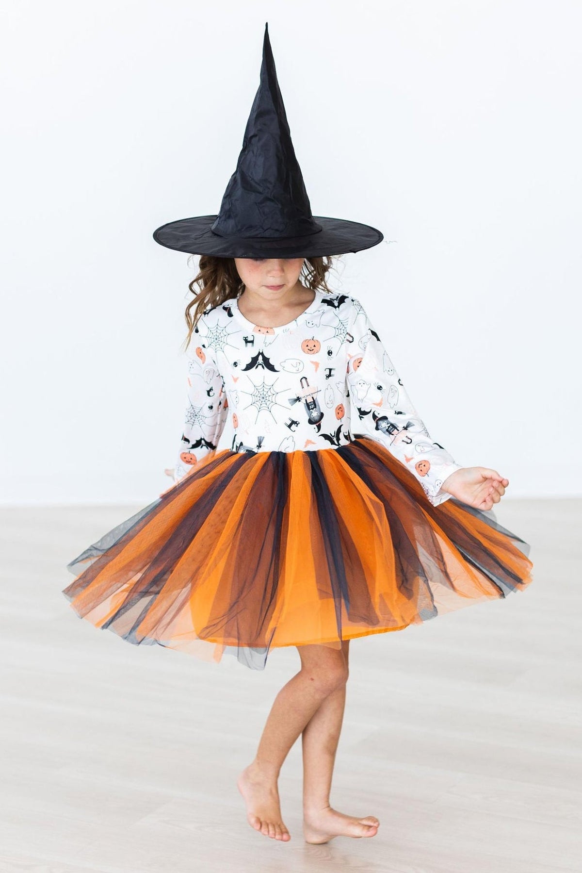 Mila & Rose Boo-tiful Halloween L/S Tutu Dress worn by a girl twirling: white print bodice with bats, ghosts, spider webs and jack-o'-lanterns over a layered orange and black tulle skirt. The black witch hat is a photo prop, not included.