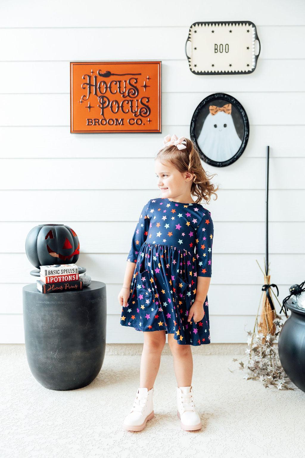 Girl walking past Halloween decorations in the Halloween Stars 3/4 Sleeve Pocket Twirl Dress, navy with multicolour stars, a gathered twirl skirt, a hair bow and white boots.