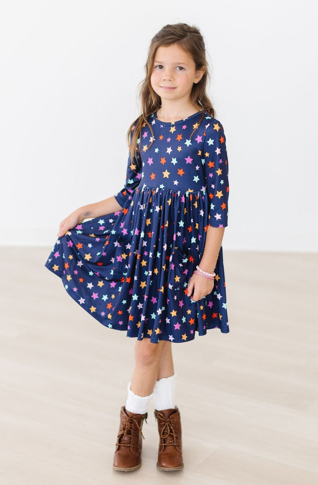 Girl holding the gathered skirt out to the side in the Halloween Stars pocket twirl dress, navy with multicolour stars, shown against a plain white background with brown lace-up boots and white socks.