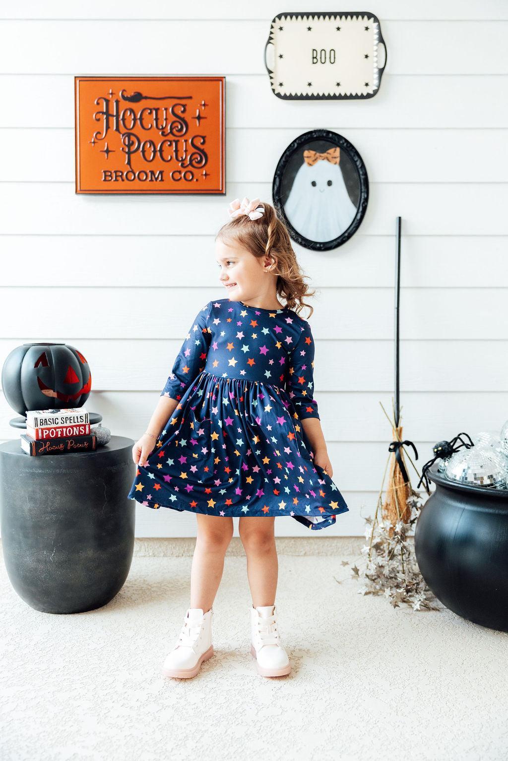 Girl turning in the Halloween Stars pocket twirl dress beside a cauldron and broom prop in a Halloween-decorated room, wearing white boots and a hair bow.