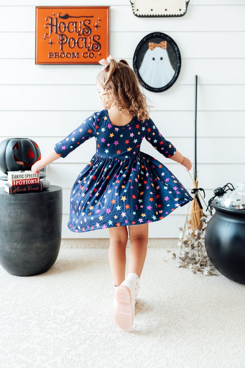 Back view of the Halloween Stars 3/4 Sleeve Pocket Twirl Dress, navy with multicolour stars, worn by a girl twirling in a Halloween-decorated room with a 'Hocus Pocus Broom Co.' sign, a ghost portrait and a cauldron.