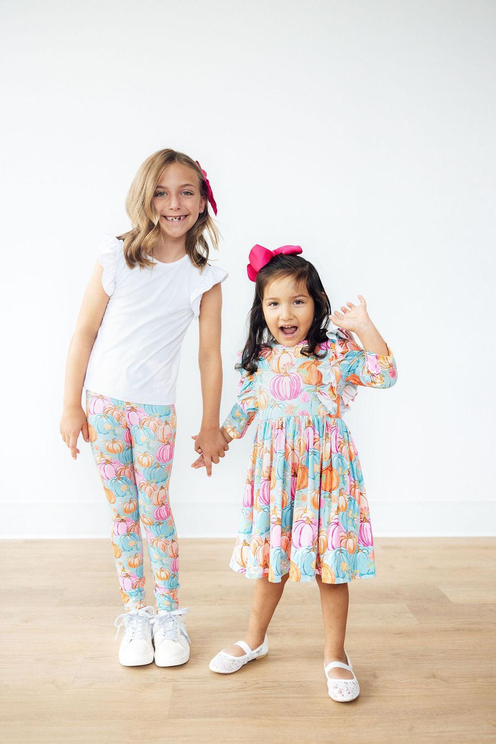 Two girls holding hands, one wearing the Preppy Pumpkins leggings with a white flutter-sleeve tee and the other in a long-sleeve twirl dress in a coordinating pumpkin print.