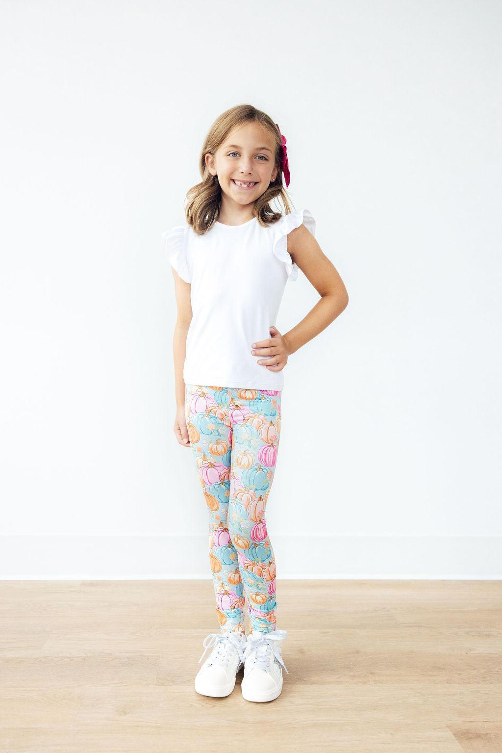 Full-length view of a smiling girl wearing the Preppy Pumpkins leggings, a white flutter-sleeve tee, white sneakers and a bright pink hair bow.