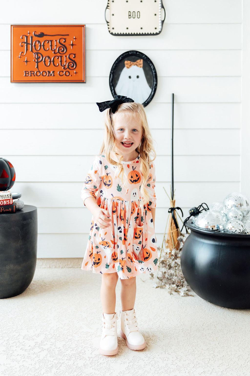 Girl in the Pumpkin Peekaboo twirl dress standing beside a cauldron in front of a Halloween wall sign, full length.