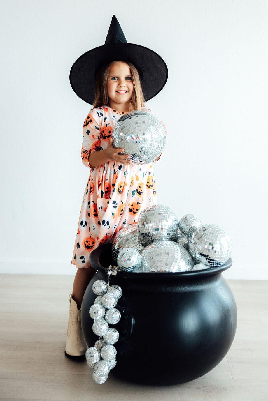 Girl in a black witch hat holding a silver jack-o'-lantern prop over a cauldron while wearing the Pumpkin Peekaboo twirl dress.