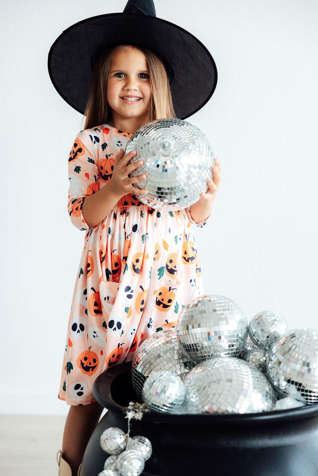 Girl in a black witch hat holding a silver pumpkin prop beside a cauldron of disco balls, wearing the Pumpkin Peekaboo twirl dress.