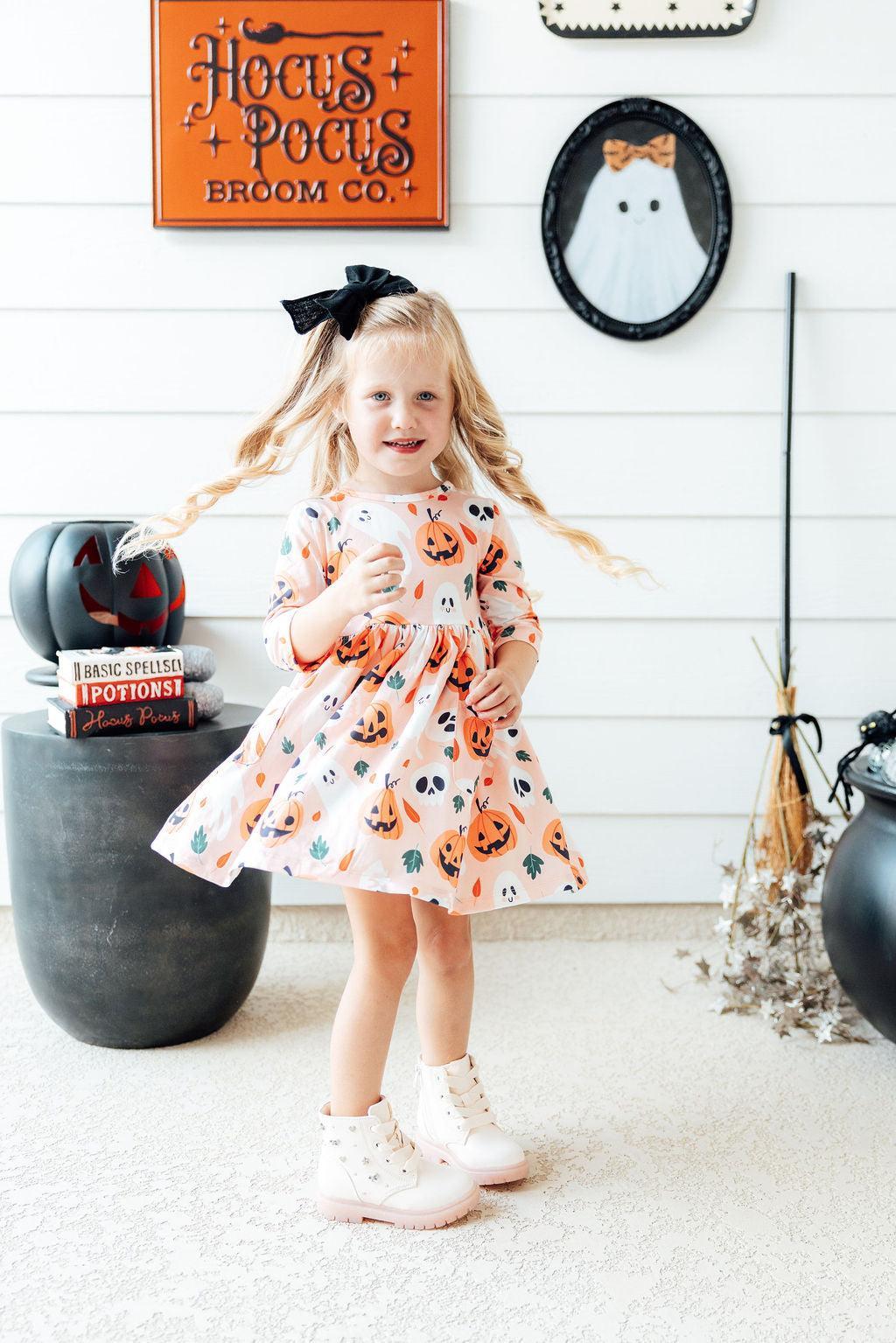 Girl wearing the Mila & Rose Pumpkin Peekaboo twirl dress, full length, in a Halloween display with cauldrons and a broom.