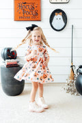 Girl wearing the Mila & Rose Pumpkin Peekaboo twirl dress, full length, in a Halloween display with cauldrons and a broom.