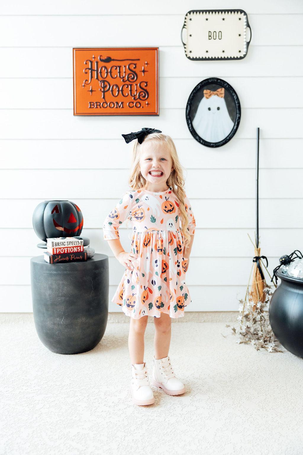 Full-length view of the Pumpkin Peekaboo twirl dress on a girl in a Halloween display with a broom and a cauldron.