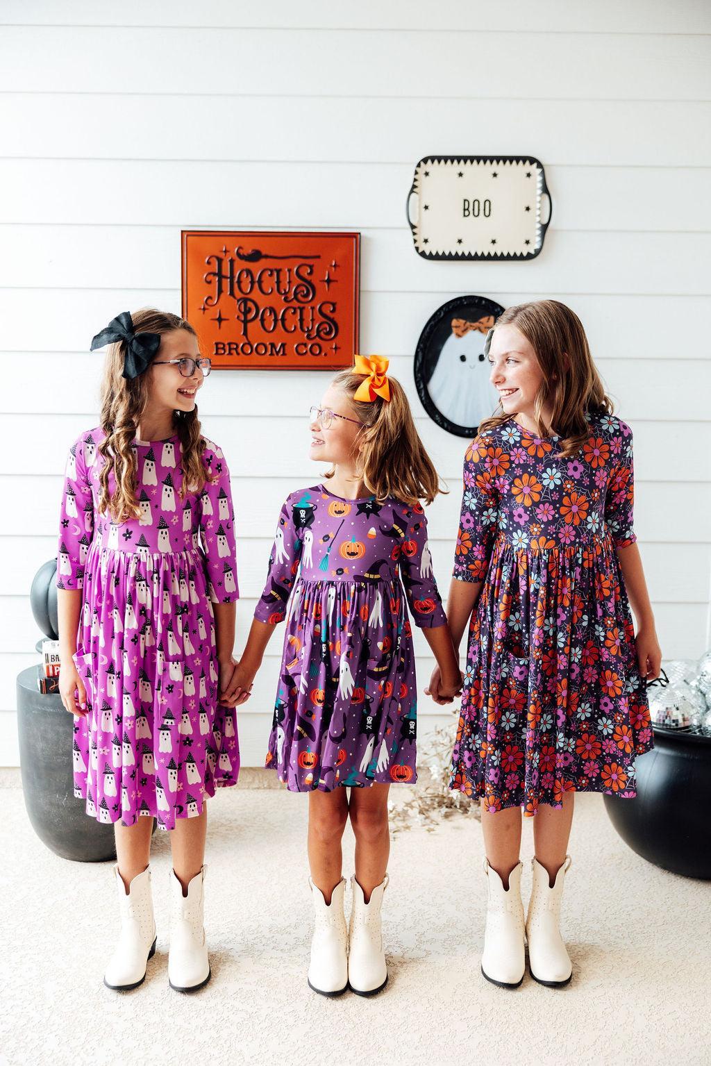 Three girls in Halloween dresses, two of them wearing the Witches Boo twirl dress, in front of a Halloween display.